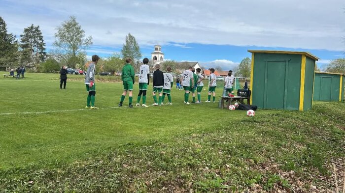 Fotbollsspelare i grönt ställ står vid sidan av en fotbollsplan, bredvid en bänk med fotbollar. En byggnad syns i bakgrunden.