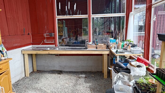 Diskbänk och hyllor i ett växthus med kringutrustning och odlingsmaterial i en lantlig miljö.