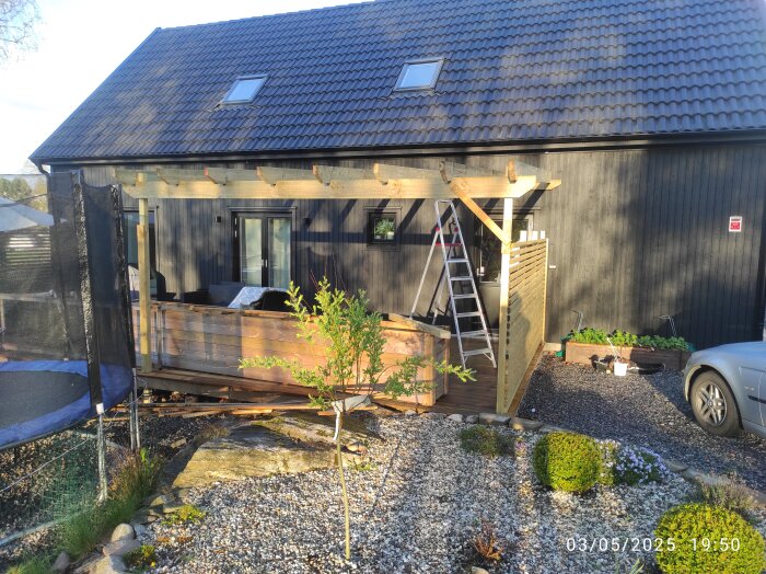 Altanbygge med träkonstruktion fast i huset. Stege lutad mot sidan, trampolin och liten buske synliga framför altanen.