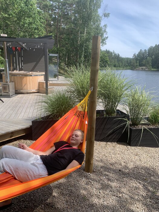Person ligger i en orange hängmatta fäst vid en stolpe bredvid en sjö med ett trädäck och en badtunna i bakgrunden.