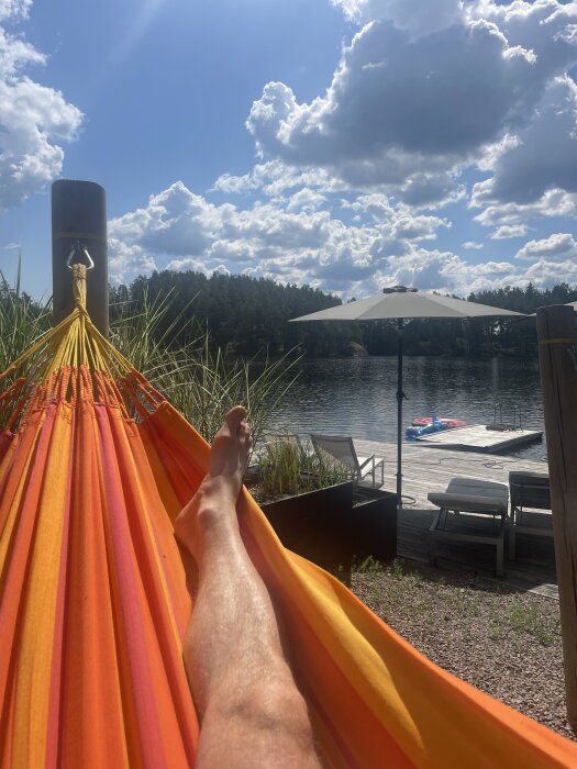 Orange hängmatta fäst i träramar med karbinhakar, vid en sjö med brygga, solstolar och parasoll under en molnig himmel.