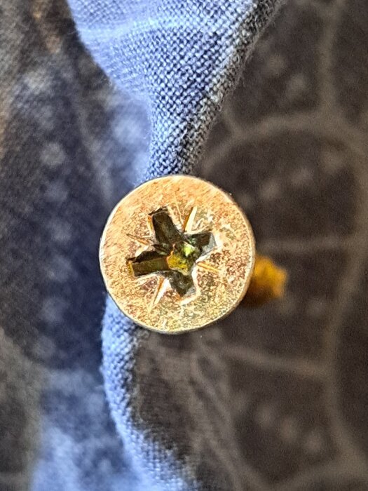 Närbild på en guldfärgad skruv med stjärnformad skruvspår inskruvad i en blå tygbakgrund.