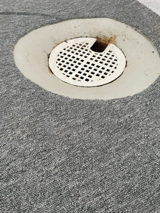 Skruvad avloppsbrunn med galler i grå matta, nerfälld i golvet.