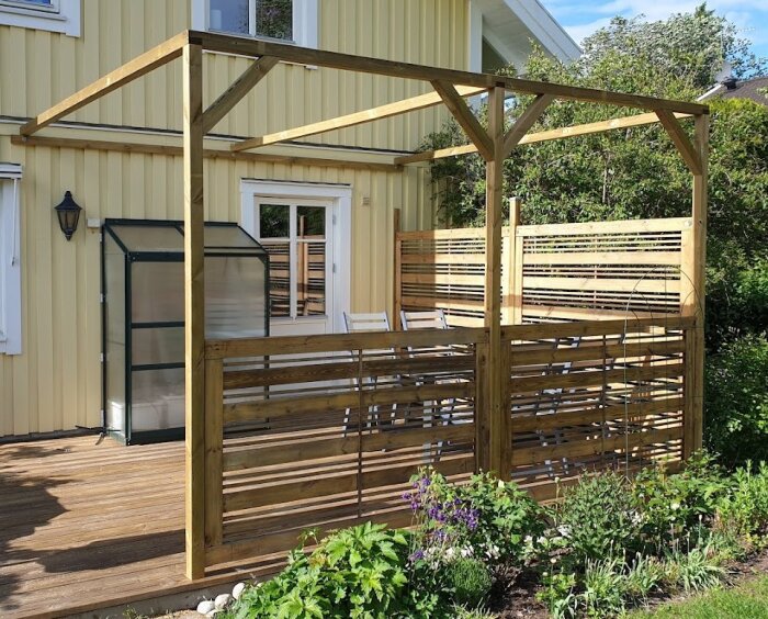 Träpergola på altan med snedsträvor, soligt väder, grönska i förgrunden, och växthus intill gulmålad vägg.