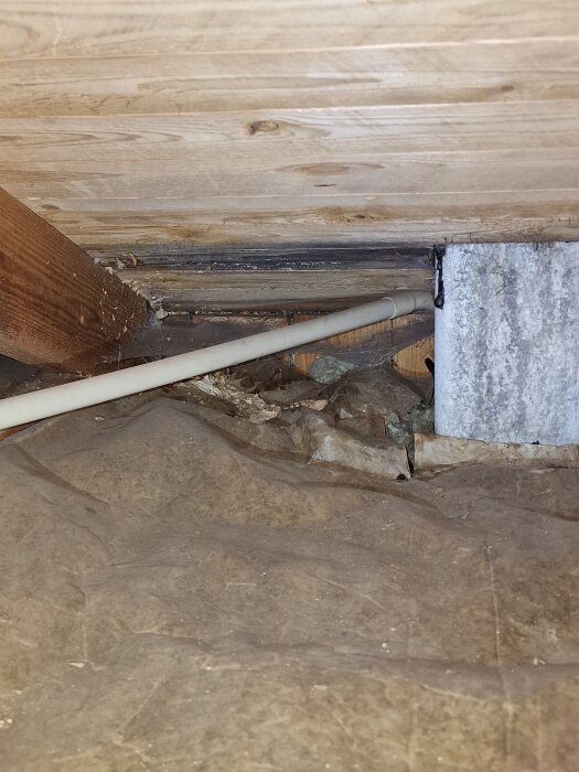 En träbalk på en vind, ett vitt rör och en möjlig fuktskadad yta intill en fläkt. Bakgrunden visar träpanel.
