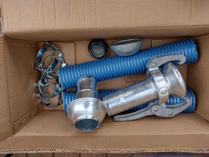 Blå korrugerad slang, metallkopplingar och klämmor i en kartong som del av ett komplettpaket för slamsugningsledning.