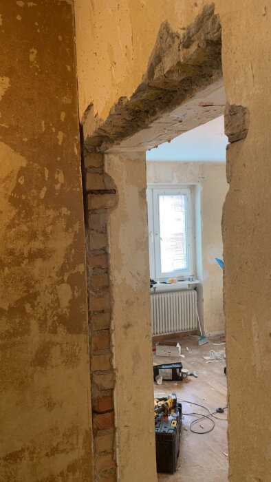 Öppning i en vägg där dörrar har tagits bort, synlig tegel och puts, rum med fönster i bakgrunden, verktyg och material på golvet.