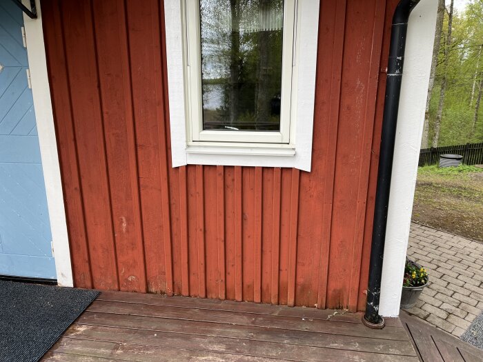 Röd stugvägg med vitmålat fönster, stående träpanel och ett tillhörande trädäck nedanför. En svart stupränna finns på sidan.