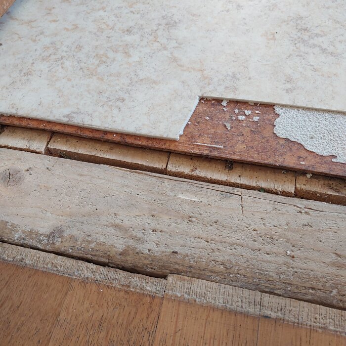 Under golvbeläggningar syns olika lager, inklusive korkgolv och parkett, vid kökströskel i ett äldre hus.