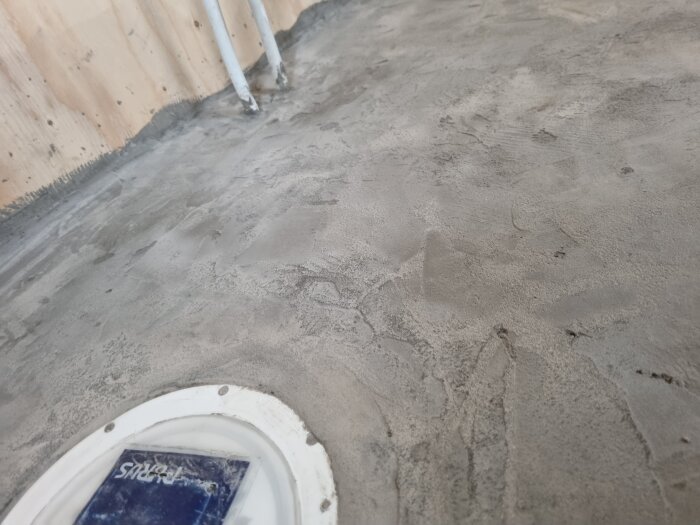 Ojämn cementgolv i badrum med vattenrör genom golvet och trävägg i bakgrunden. Rund vit inspektionslucka i förgrunden.
