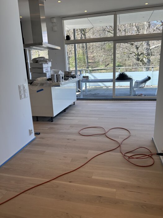 Ljust vardagsrum med stora fönster, ljus trägolv, köksö täckt med färgförpackningar och röd förlängningssladd på golvet.