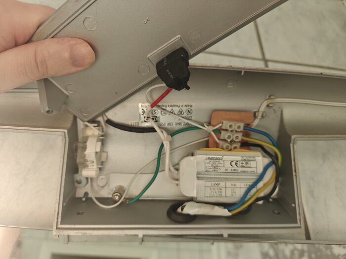 Inuti en väggbadrumslampa med bortskruvad glimtändare och blottade kablar samt komponenter, inklusive lysrörsballast.