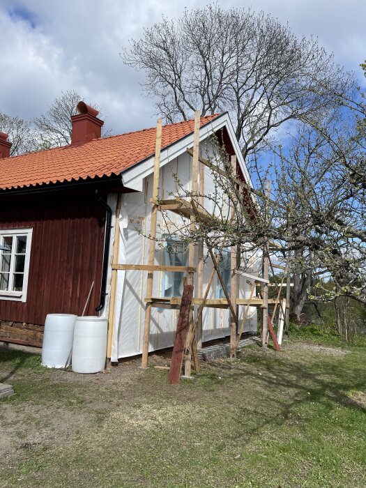Röd träbyggnad under renovering med ställning, vindduk och träd utan löv i bakgrunden.