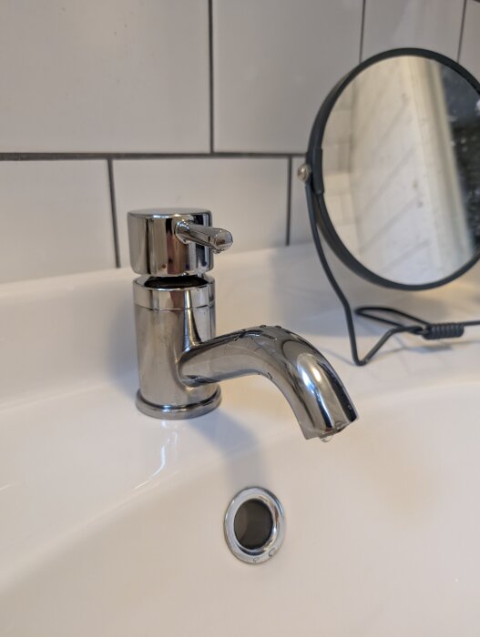 Närbild på en kromad blandare installerad på ett vitt handfat, med en droppe vatten på pipen, och en spegel i bakgrunden.