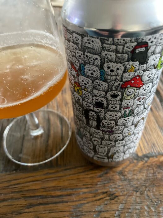 En ölburk med tecknade figurer står bredvid ett glas med bärnstensfärgad dryck på ett träbord.