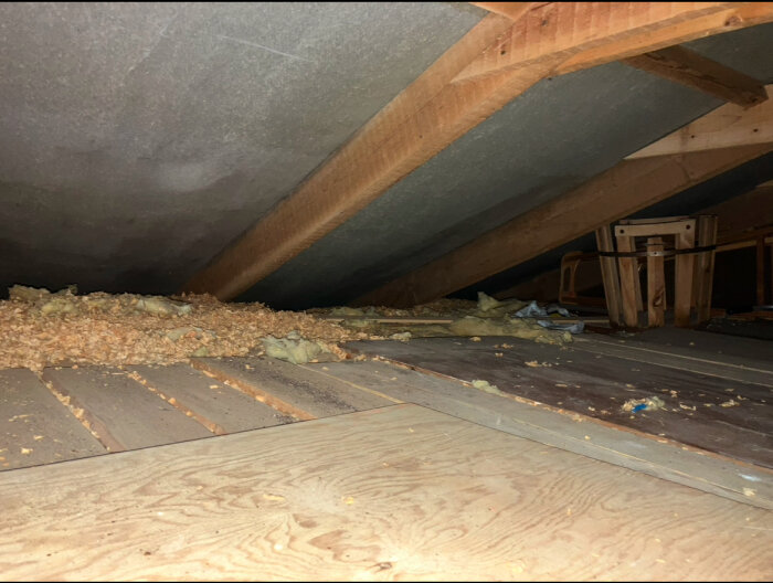Vindutrymme med grått underlagstak, träbjälkar och spån. Isolering och bråte syns på golvet.