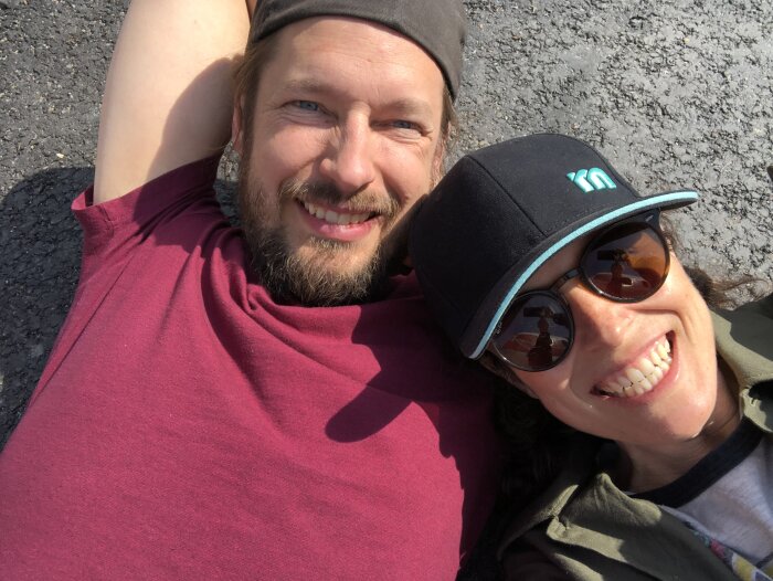 Två personer som ler ligger på marken. Den ena bär keps och solglasögon. Soligt väder.
