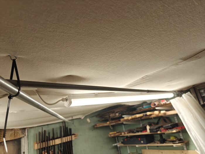 Tak med en upphängd metallstång och ett lysrör ovanför hyllor med verktyg i en verkstadsmiljö.