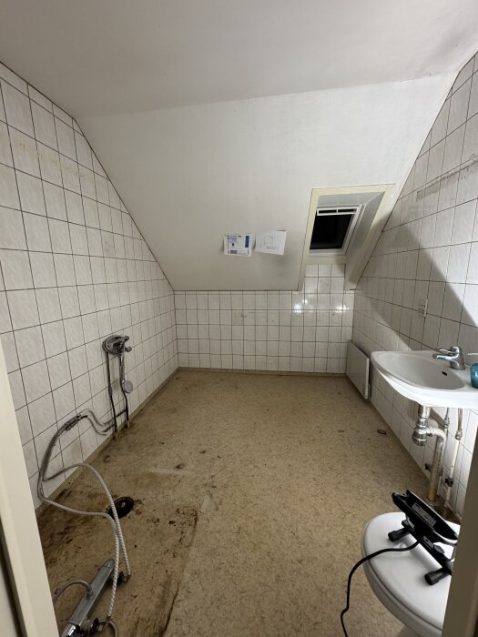 Tomt badrum med kaklade väggar, borttaget badkar och dusch, handfat kvar. Kopplingsslangar på golvet, öppet takfönster.