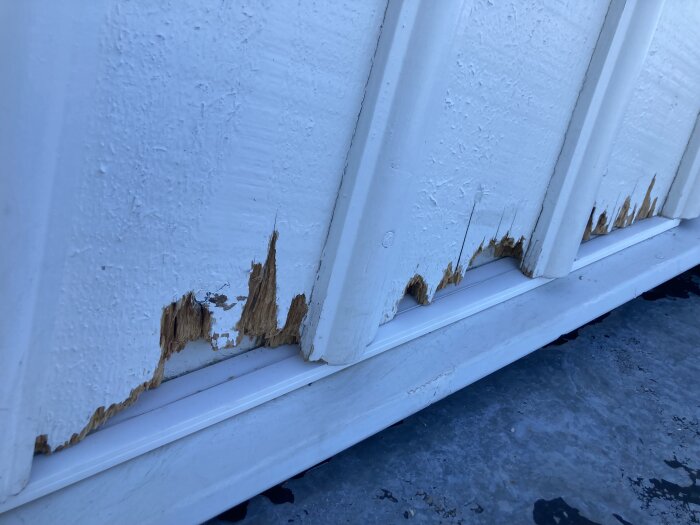 Skadad vitmålad träfasad med synliga spacklingsområden och flagnad färg på ändträ.