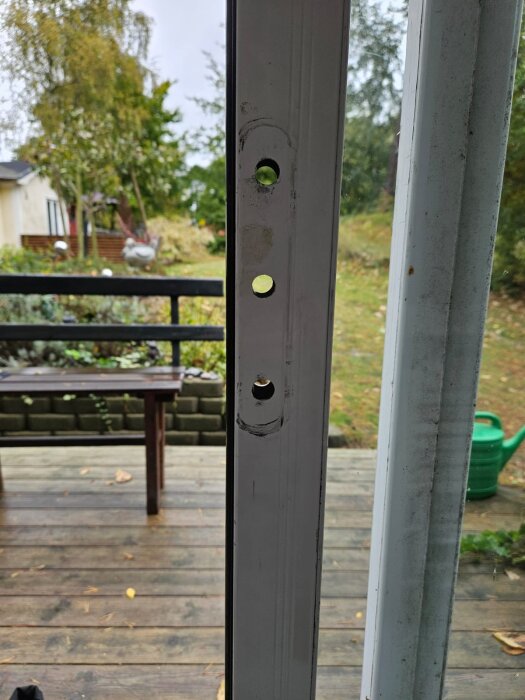 Låsets urtag på en skjutdörr av metall med utsikt över en träveranda och trädgård i bakgrunden.