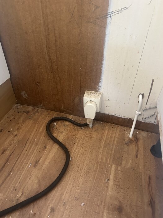 En vit Perilexkontakt på väggen med en svart elkabel som ligger på ett trägolv i en sommarstuga.