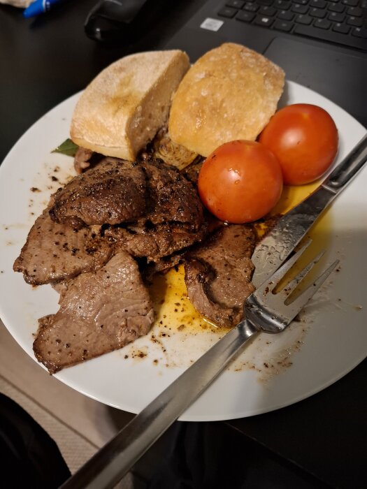Tallrik med lövbiff, två hela tomater, brödskivor och en gaffel, placerad vid en laptop.
