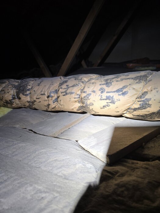 Ventilationsrör med synlig påväxt på isoleringen i mörk vindsmiljö, upplyst av ficklampa.