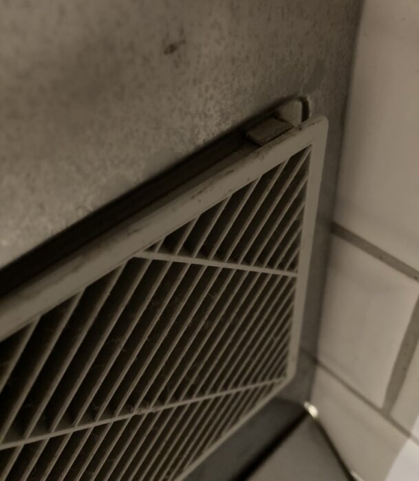 Närbild av ett lutande ventilationsgaller, del av en fläkt, där en sektion är snett monterad. Bakgrund med kakel och metall.