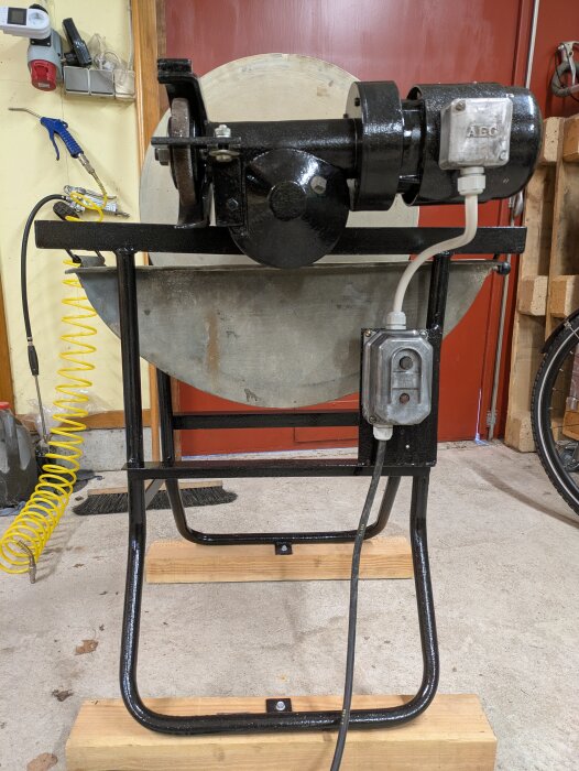 Slipmaskin återuppbyggd, nyoljefylld och placerad på träblock i ett garage med röd dörr och verktyg runtom.