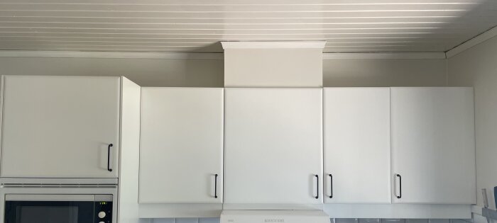 Kök med vita överskåp och ett gap mellan skåpen och taket. Skåpens beslag är svarta och taket är av vitmålad träpanel.