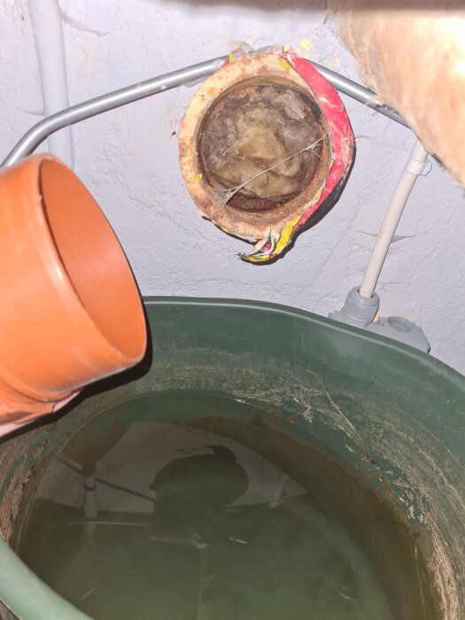 Pluggat rör i källare med röd tejp runt öppningen, orange rör bredvid med anslutning till en grön tank med vatten.