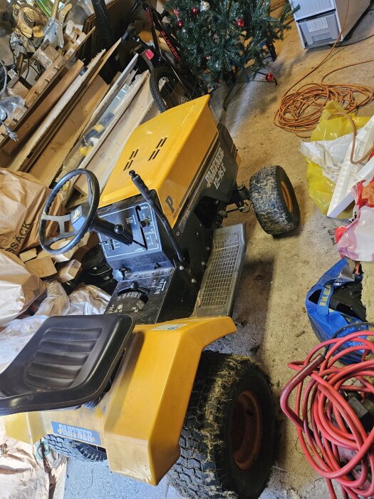Gul Partner 12-95E åkgräsklippare i ett garage, omgiven av kablar och verktyg, med en cykel och en julgran i bakgrunden.