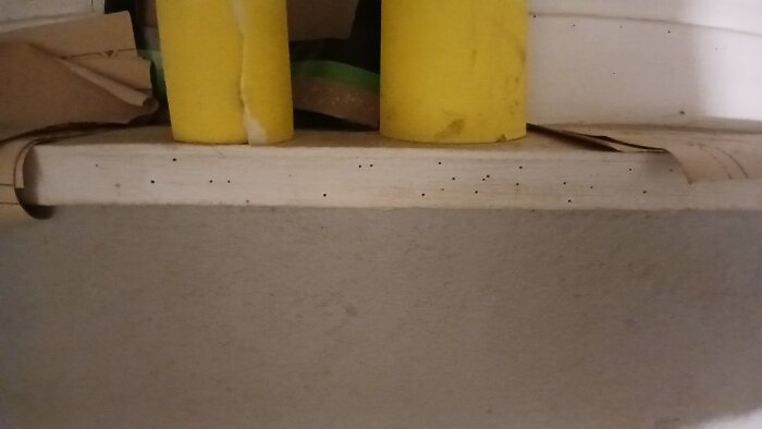 Träbräda med små hål och uppsamlat träspån nedanför, troligen orsakat av insekter; gul föremål i bakgrunden.