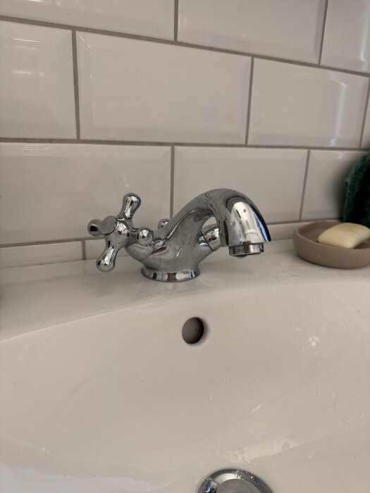 Kromad tvättställsblandare med korshandtag mot vit kakelvägg.