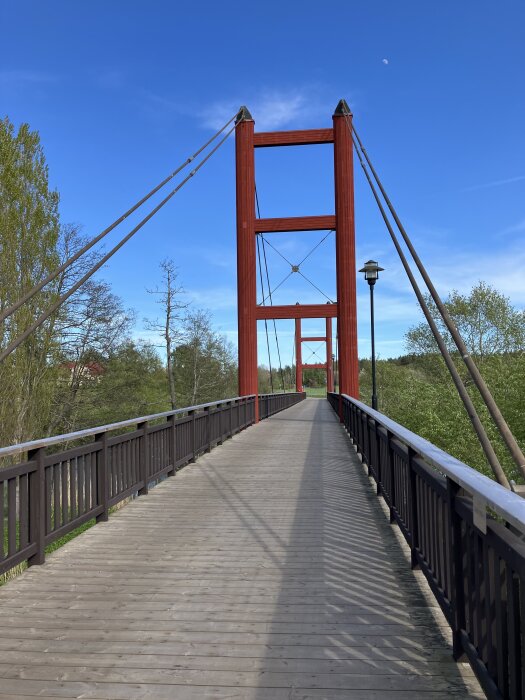 Röd hängbro över grönskande landskap med blå himmel i bakgrunden.