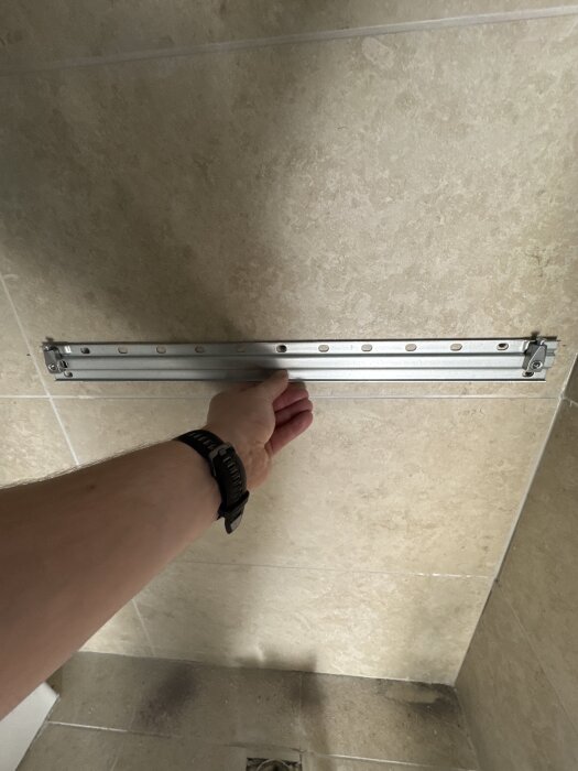 En hand håller upp en monteringsskena mot en vägg med beige kakel, förberedelse för att montera en kommod.