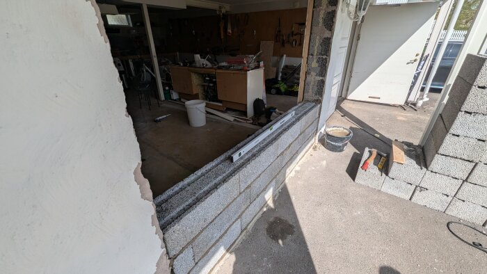 Byggarbetsplats med delvis murad vägg av lecablock och verktyg i ett garage. Vattenpass ligger på väggen och byggmaterial syns runtomkring.