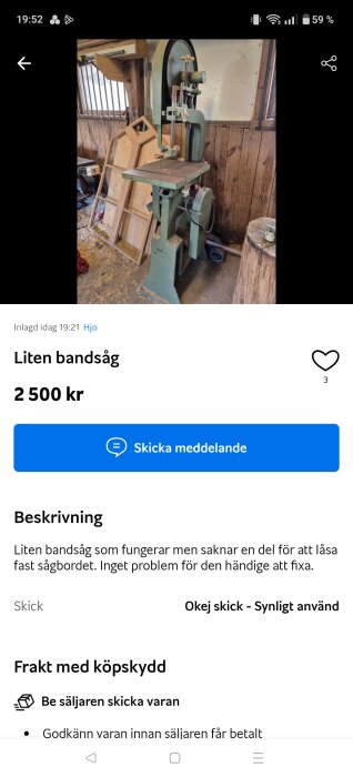 Grön bandsåg i ett träverkstad, omgiven av träramar. Sågen saknar en del för att låsa sågbordet. Sågspån är synliga på golvet.
