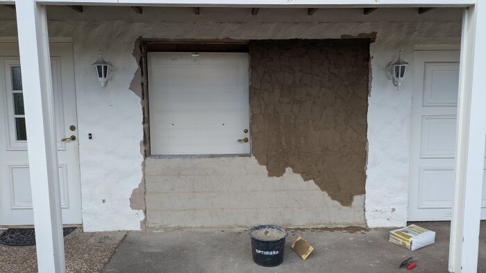 Vägg under renovering med lecablock, putsad yta och verktyg framför. Gamla garageporten täcker del av öppningen.