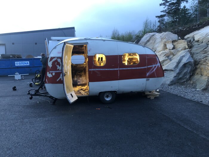 En gammal husvagn under renovering, med öppen dörr och upplyst interiör, står parkerad på asfalterat område i skymningen.