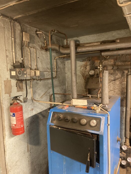 Panna med rörsystem i källare, flertalet rör och ventiler synliga med en röd brandsläckare monterad på väggen bredvid.