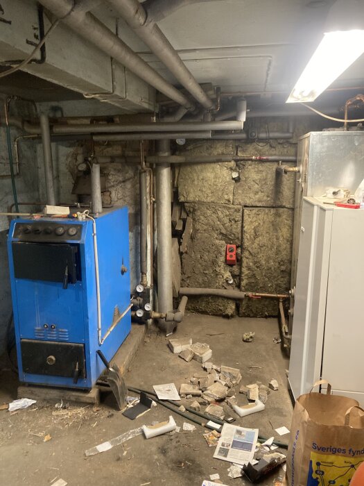 Pannrum med blå värmepanna, många rör i taket, och skräp, tidningar samt stenblock på golvet.