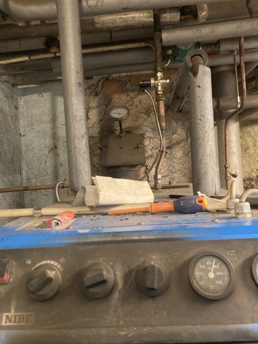 En äldre pannanläggning med synliga rör och en temperaturskala. Det ligger verktyg som en tändare och skruvmejsel ovanpå pannan.