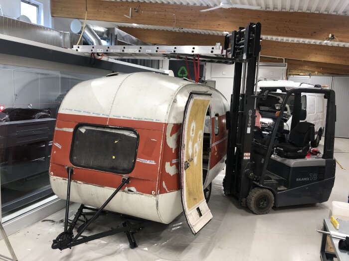 Husvagn på verkstad, stödd av en gaffeltruck med öppen dörr, i pågående renoveringsprojekt.