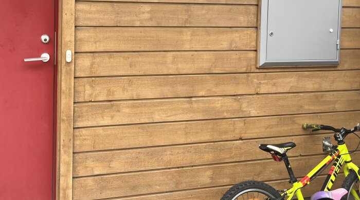 Träpanel i brun nyans med röd dörr och en gul cykel i förgrunden.