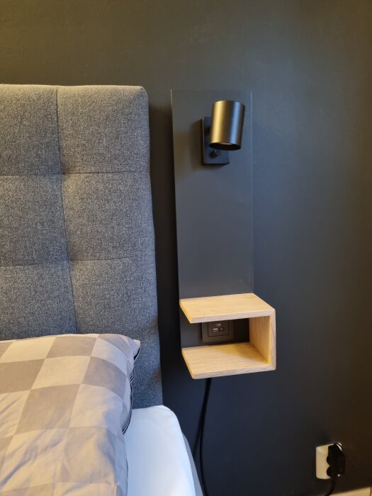 Grå sänggavel med vägglampa och hylla i trä, eluttag nedanför, mot mörk vägg.