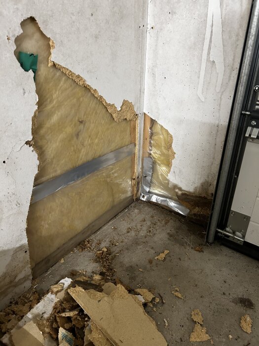 Riven gipsvägg visar isolering och en del utan skum/plastduk, med fuktig träregel synlig. Brädrester och skräp på golvet.
