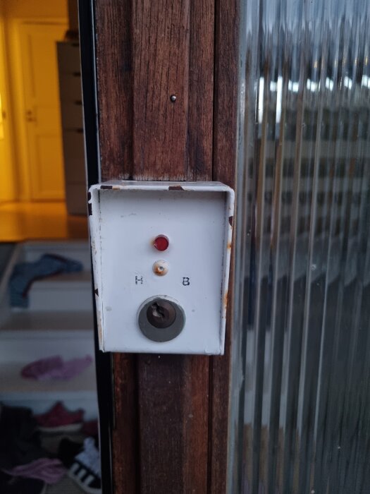 En äldre vit apparat med en röd och en vit knapp samt ett nyckelhål, monterad på en träkarm vid en ytterdörr.