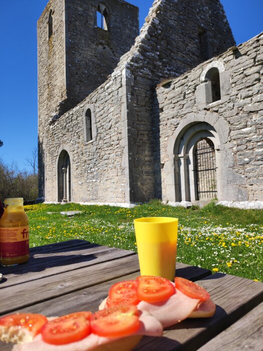 Ödekyrka i bakgrunden med en picknick på ett träbord i förgrunden, bröd med tomat och dryck.